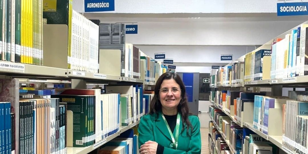 Professora Elizabeth Giron Cima em frente a uma estante de livros em um ambiente acadêmico.