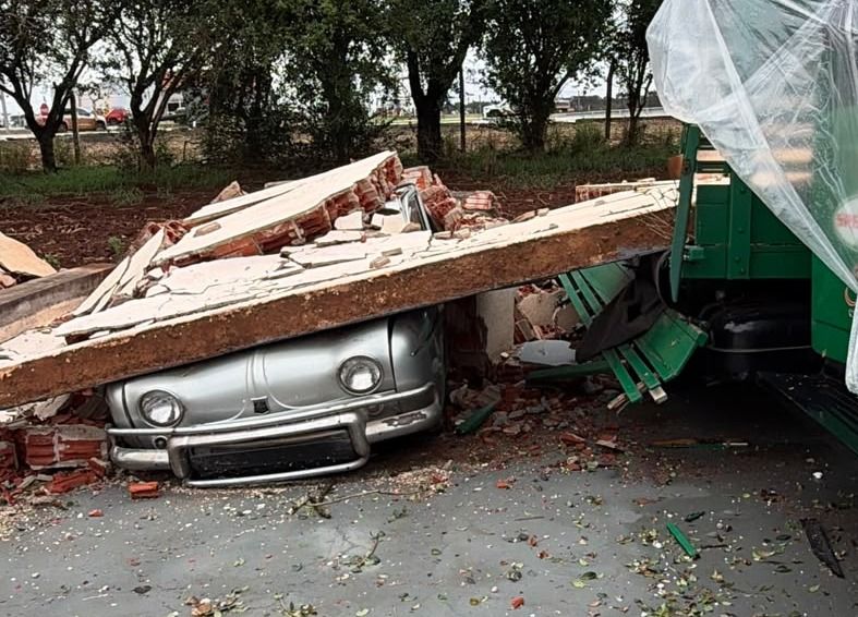 Estrutura de um barracão destruída após forte temporal, com destroços espalhados sobre carros clássicos e caminhões danificados.