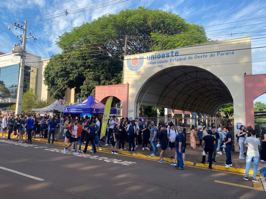 Fachada do campus da Unioeste em Toledo, Paraná, com estudantes entrando e saindo do prédio.